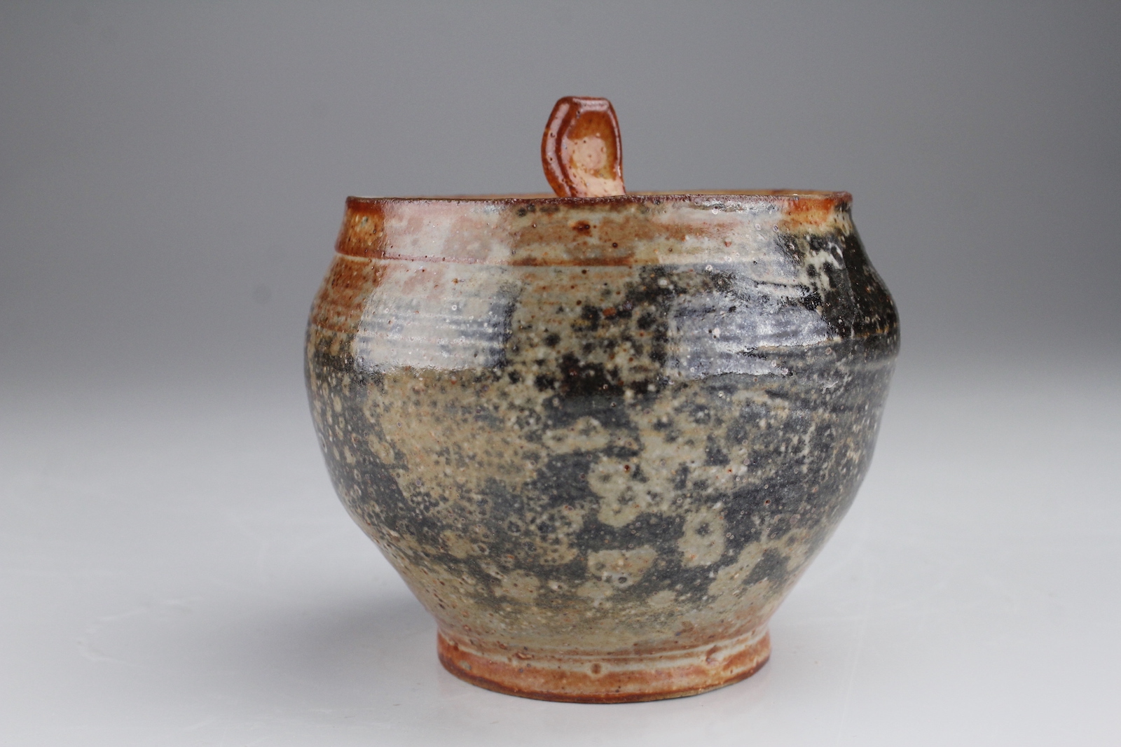 a jar with a small foot balooning outwared to a wide top. The lid has a nob that looks like a rectangle gently pressed inwards by a fingertip. The glaze is red with splotches of black and greyish brown covering much of the surface.