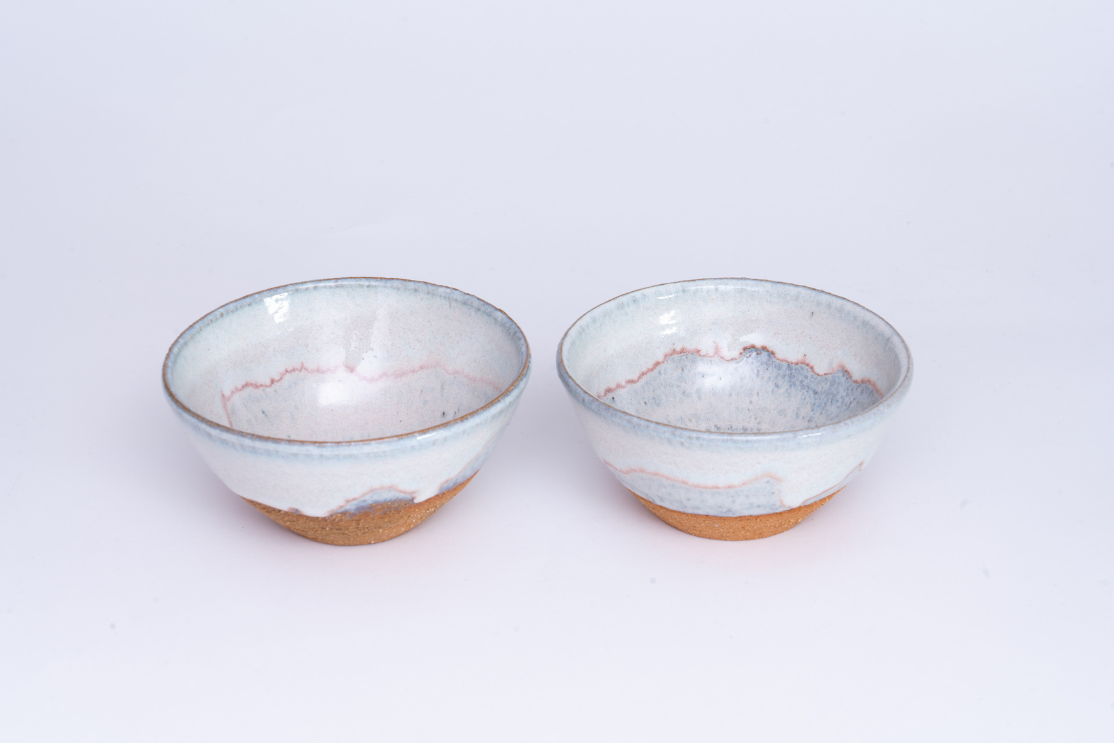 two deep bowls with no foot covered in a white, glassy glaze
            that breaks blue at the edges with a faint wavy line of pink inside