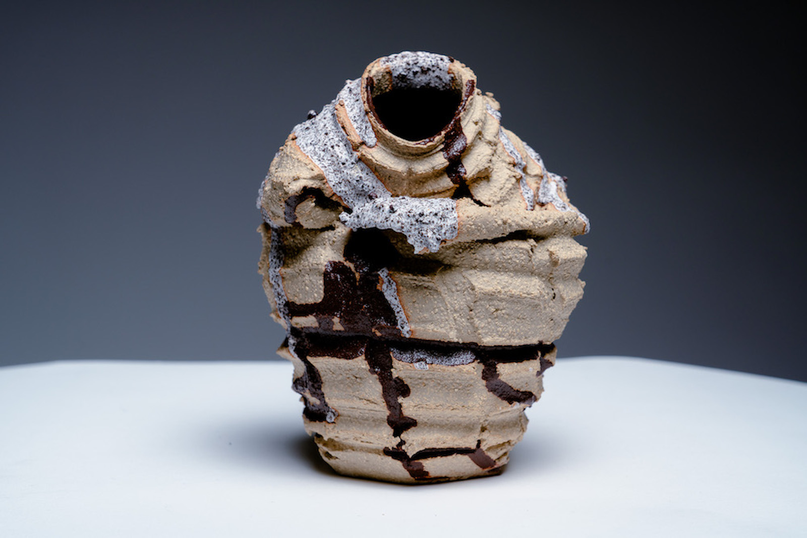 a vase with rough rectangular cuts 
            all over it's surface. It has a darker brown and white glaze running through its grooves.
            The top of the pot is flopped over so the neck faces the camera.