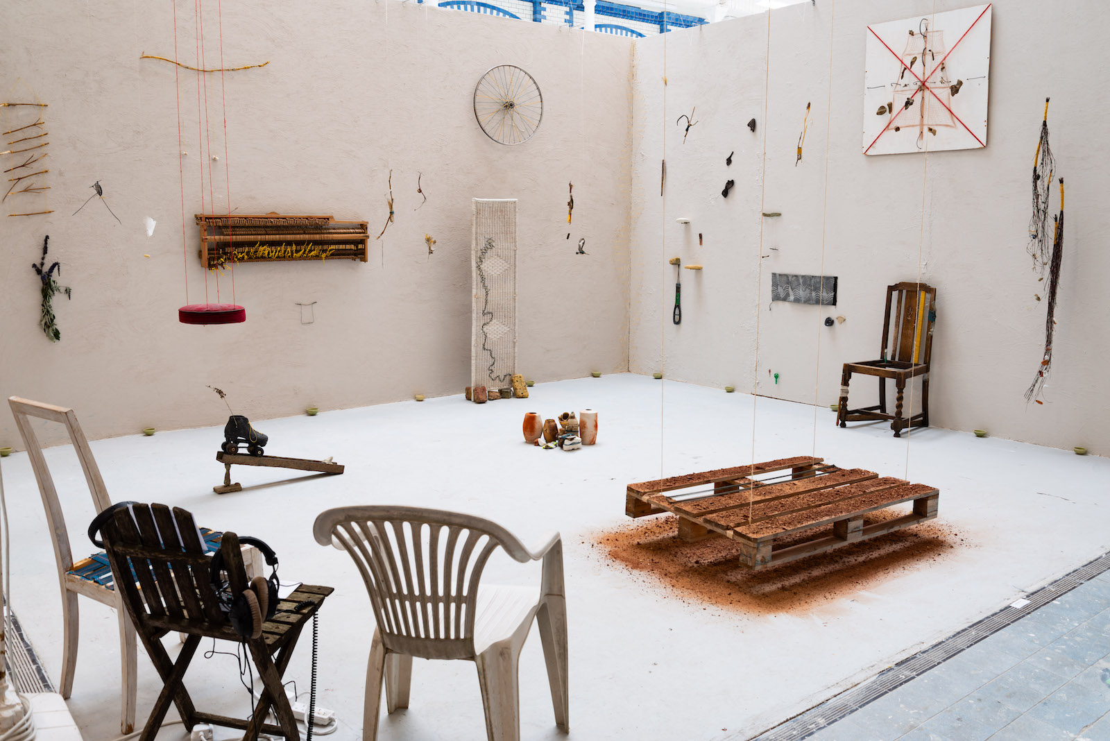 a view diagonally showing the entire installation space
        there are three chairs in the forground on which headphones are draped. 
        A pallet is suspended just above the ground and is covered in brick dust.
        a roller skate sits on top of a U-beam some sort of stone propping it up 
        at one end. A collection of brownish grey pots, plastic bottles, bits 
        of chalk and other rubbish items are clustered in the midground. Many of 
        these items have a belt of yarn wrapped around them. A chair sits 
        against one wall, its seat missing, thread wrapped around one of its legs 
        and part of its back. A wicker room partition stands near one wall
        with the Thames embroidered through the wicker