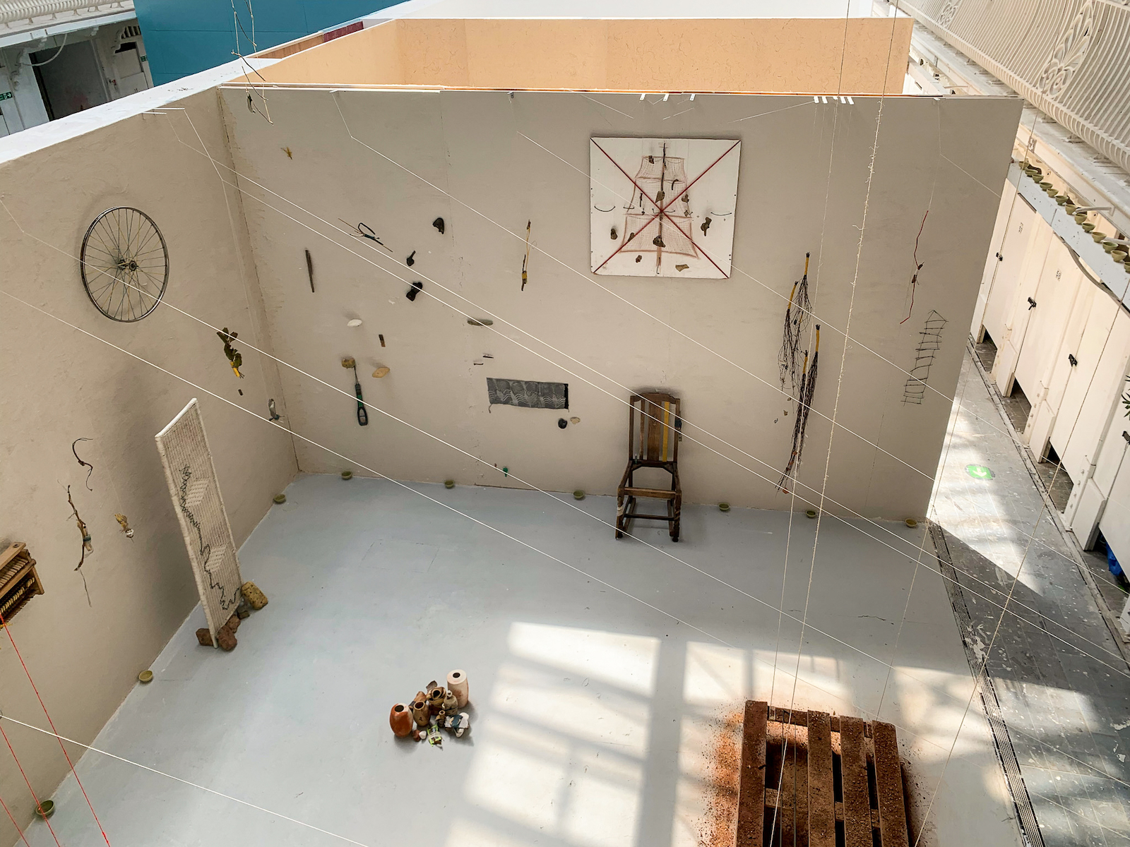 a view towards the pieces <em>Chair<em/> and <Crimson Tide<em/>. From above, the light also catches
            the light of strings webbing out towards both walls from the balcony. These strings then hold the various
            small sculptures suspended in front of the walls.