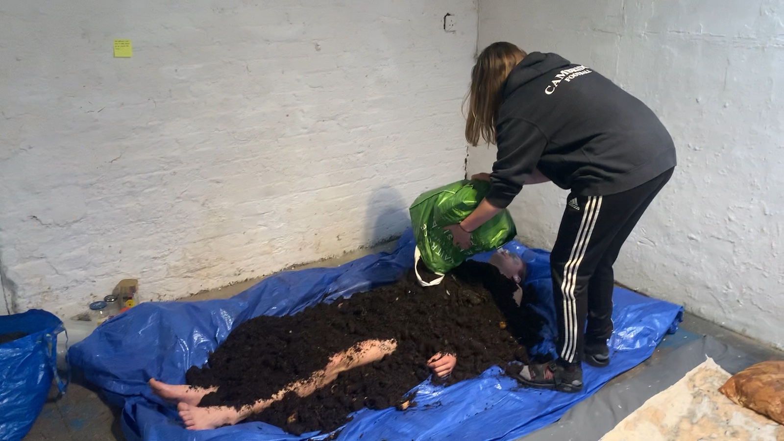 me, lying on a blue tarp with a friend pouring compost over 
            my mainly covered body.