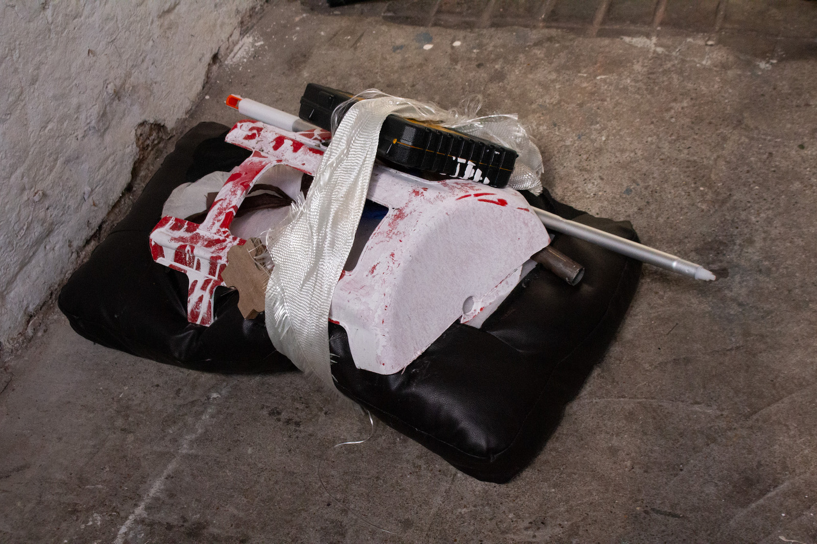 a leather couch cussion, red stool with white paint on it, plastic tool box, and half a mop handle wrapped around with a tattered white 
            strong fiberglass towing strap wrapped around them.