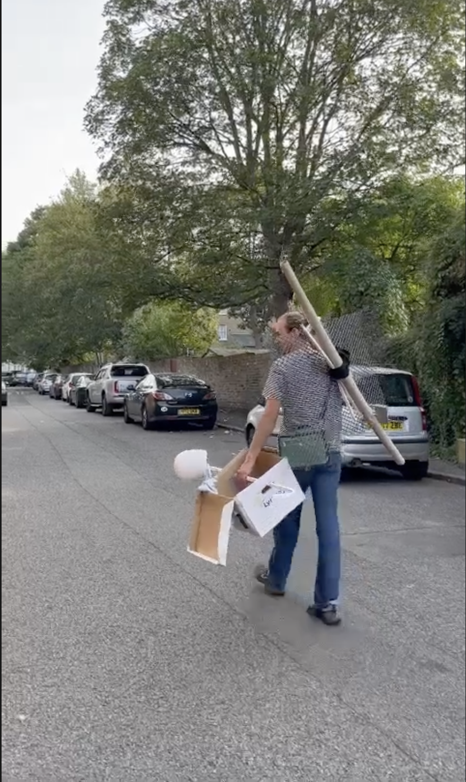 an image of the artist from the back walking 
            down a street with a metal grate with rods attached to it with a piece of black cloth in one hand 
            and a filing box with a curtain rod through the handles which has a mannequin head on one end in the other.