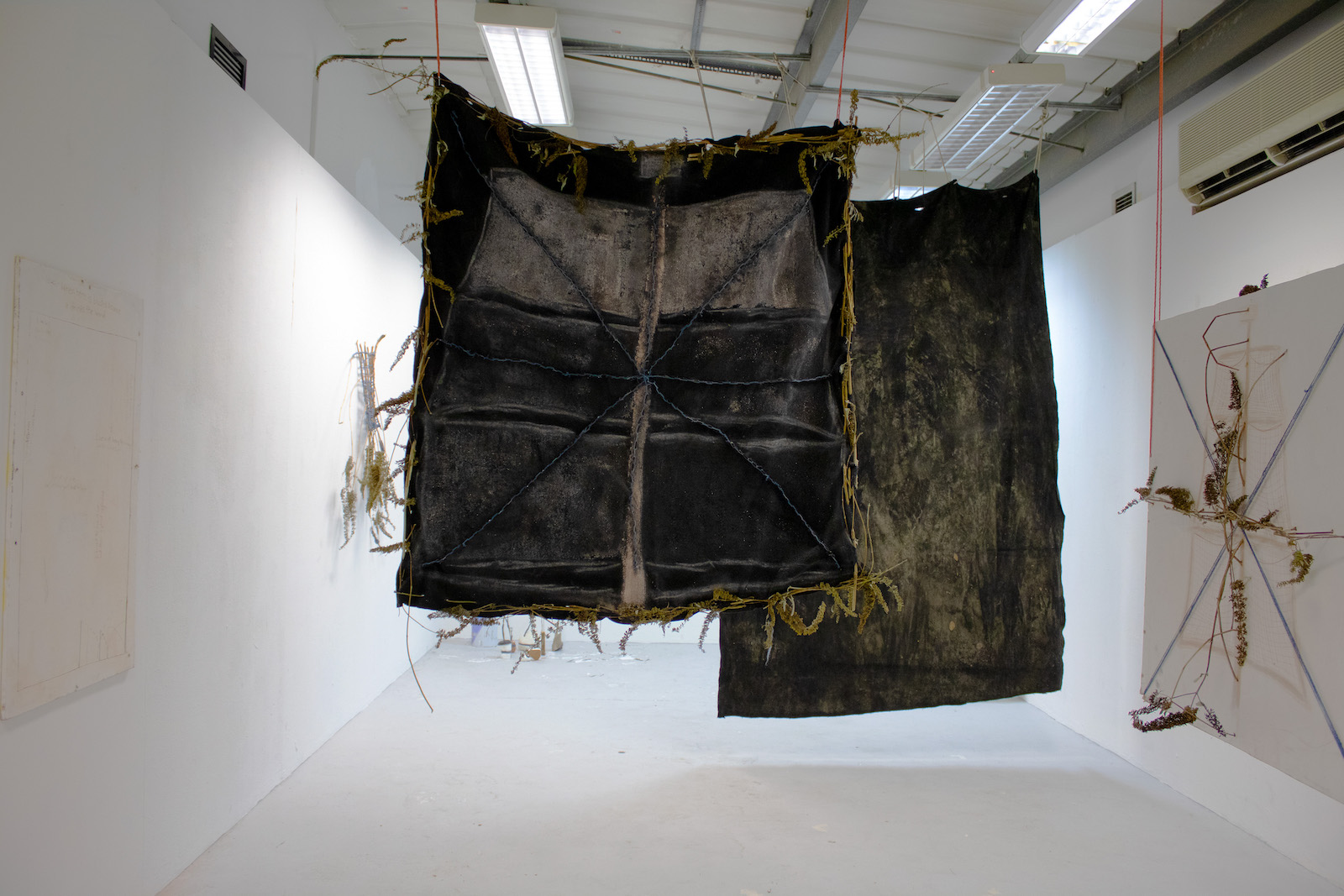 a long room in which a black fabric is hung with a grey
            pink sailing ship mast on it. The fabric is framed by butterfly 
            bush. Behind it is another black fabric covered in mud.