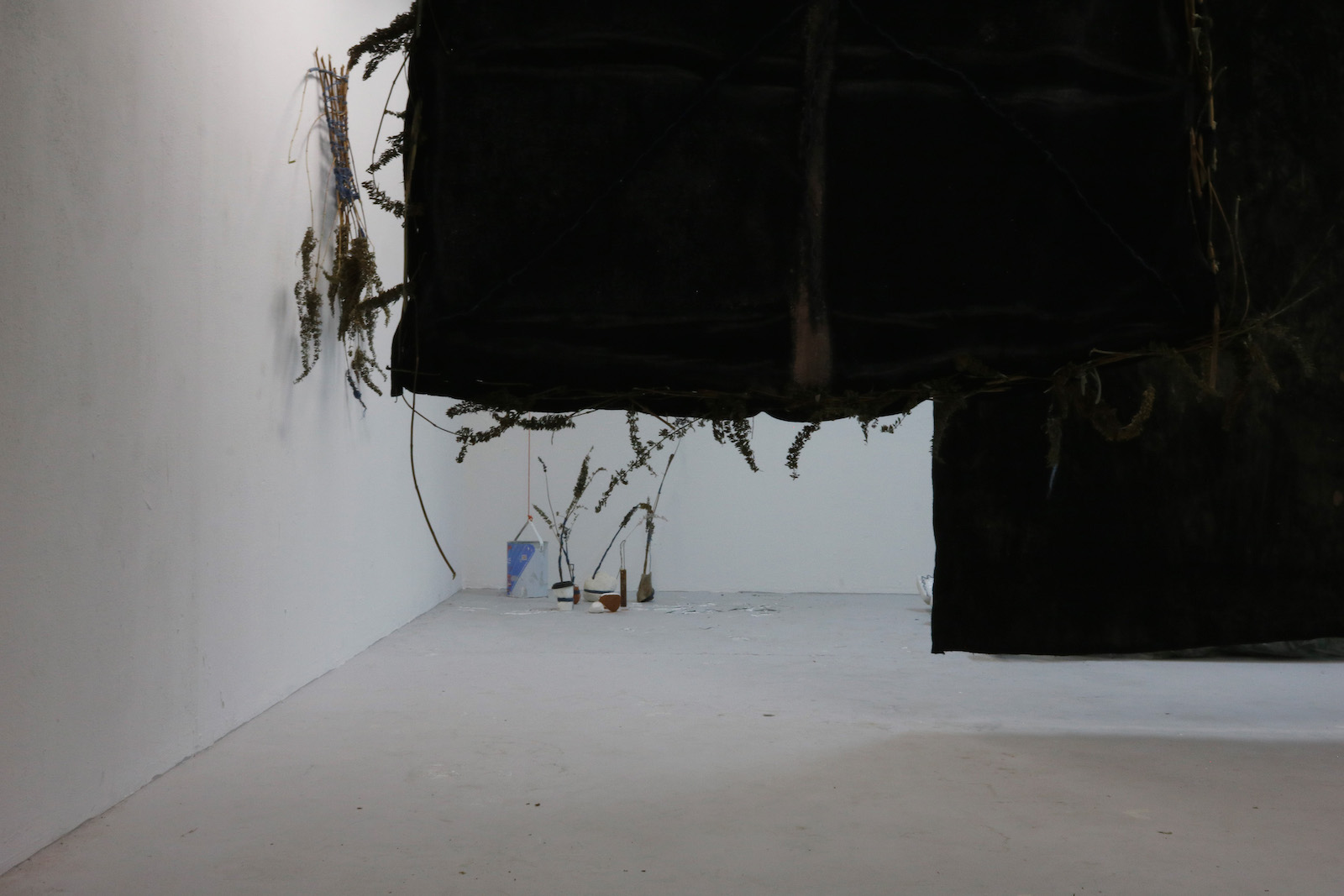 a view below the black fabric pieces in which a small cluster of objects
            including a piece of chalk wrapped in blue yarn, a paint can, and various bits of
            bone and brick used as vases for butterfly bush are visible.