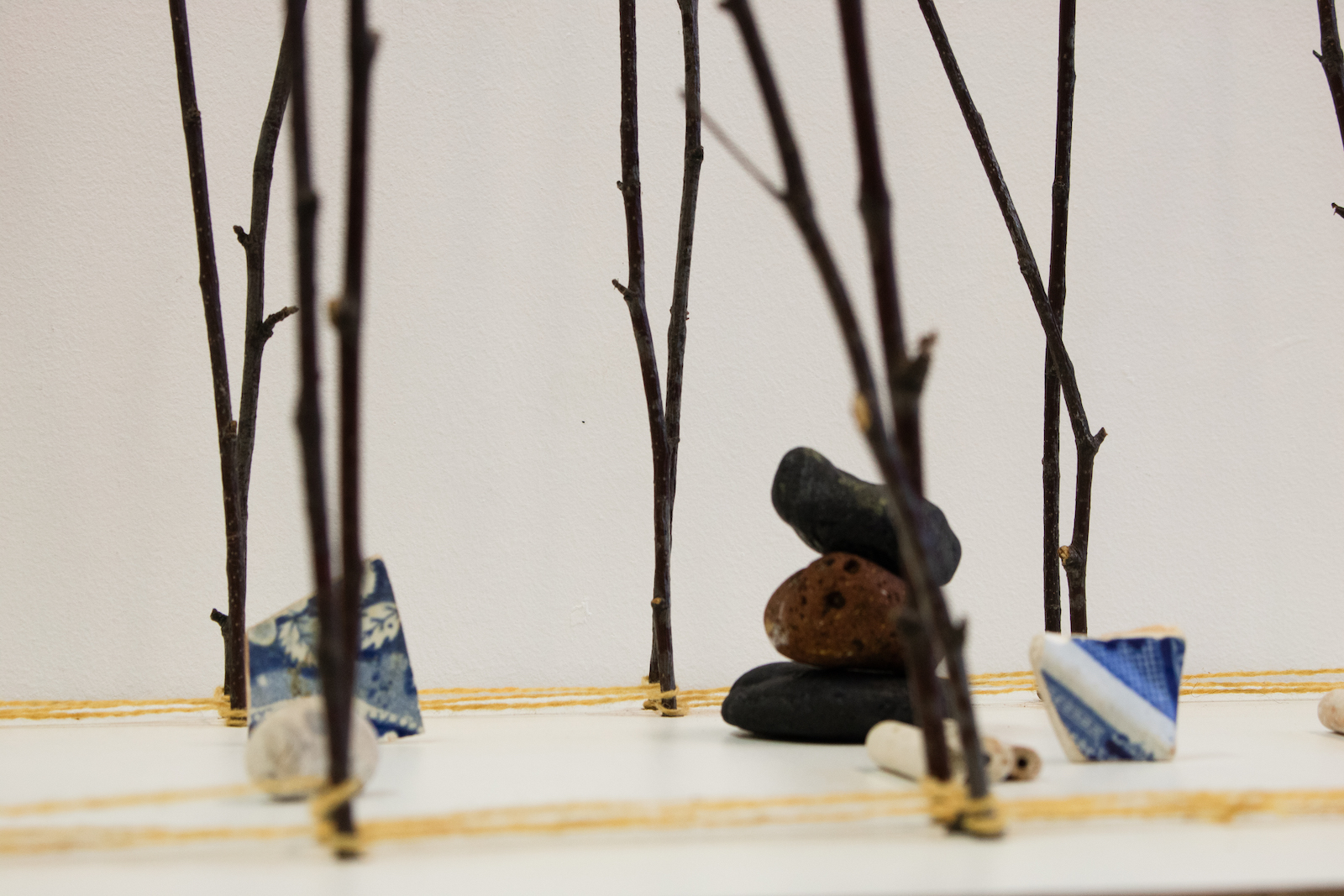 a closeup of part of the sculpture showing a pile of pebbles of brick and coal beside fragments of blue and white ceramic. These objects are seen between two lines of vertical twigs tied together with string.