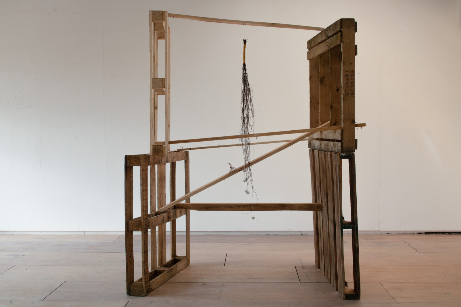 pallets stacked on top of each other connected by various pieces of wood.
           On one of these pieces at a steep angle there is a ceramic figure of a child.
           There is a bundle of birch branches with bits of clay tied into it hanging from the top of the pallets